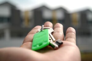 Das Bild zeigt eine ausgestreckte Hand mit einem Wohnungsschlüssel und Wohnhäusern im Hintergrund.
